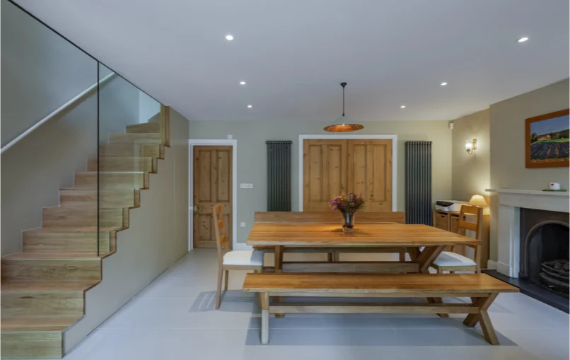 This image depicts the downstairs living space of the house. On the left, you can find the stairs that are leading to the upper floor. In the center, there is a bench with with a fire place on the right. In the back, you can find 2 doors; one leading to the rest of the house and the other to a utilities cabernet.