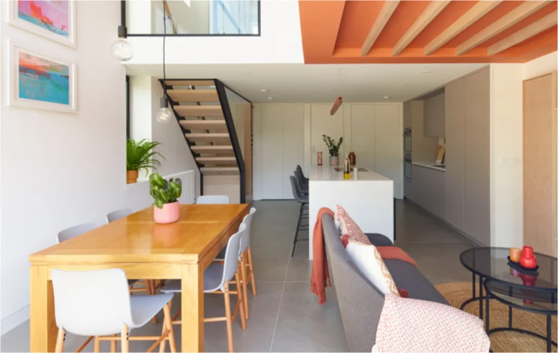 This image depicts an open plan living room, showing off the inside of the cube house. You can see the kitchens island, the sofa, and dining table with stairs in the back left leading to the top floor.