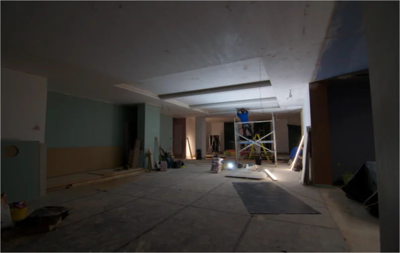 This image depicts the whole basement under construction with plaster boards on the walls and ceilings and an electrician mounting LED strips to the ceiling.